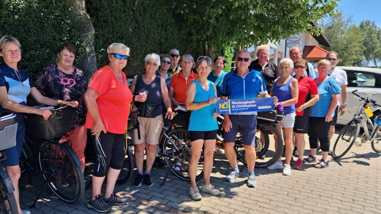 Eine Gruppe von Menschen in Fahrradausrüstung steht draußen und hält ein blaues Schild mit NO darauf. Sie posieren mit ihren Fahrrädern.