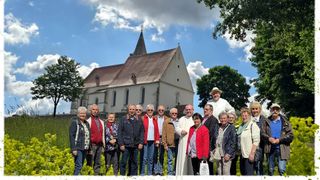 Eine Gruppe von Menschen steht vor einer Kirche, einige tragen eine Brille und Jacken, hinter ihnen steht ein Mann in einer weißen Robe und einem Hut.