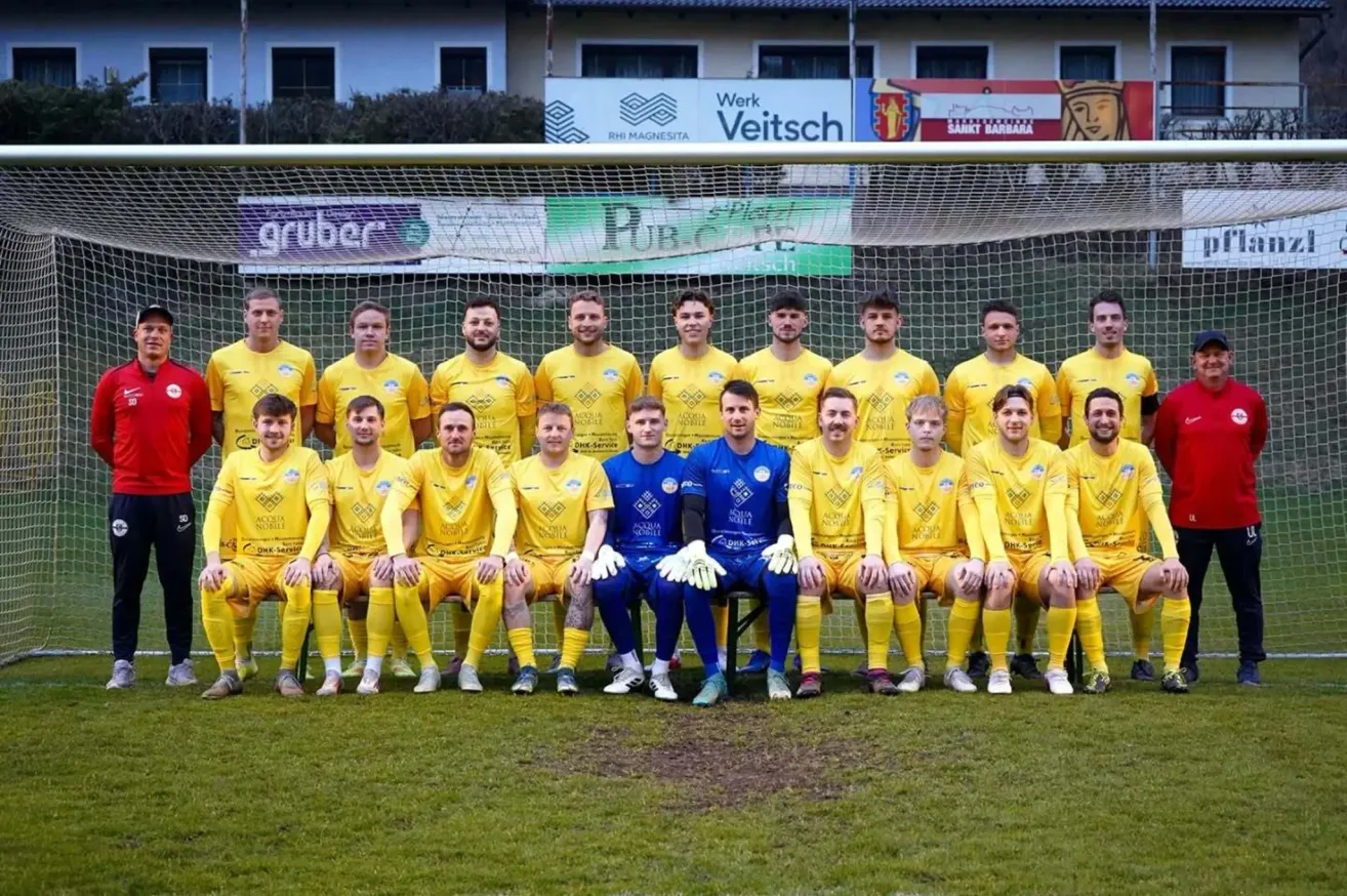 Eine Fußballmannschaft in gelben Trikots posiert für ein Foto vor einem Tor, wobei zwei Spieler in blauen Trikots in der Mitte sitzen.