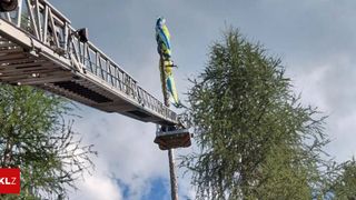Ein Feuerwehrmann in einem Gurt klettert eine Leiter zu einer Flagge auf einem Pfosten hinauf, wobei er eine Leiterverlängerung benutzt. Der Himmel ist bewölkt.