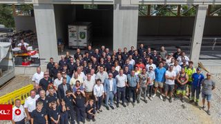 Eine Gruppe von Menschen in Uniform steht vor einem Gebäude mit einem LKW im Hintergrund. Sie posieren für ein Foto.