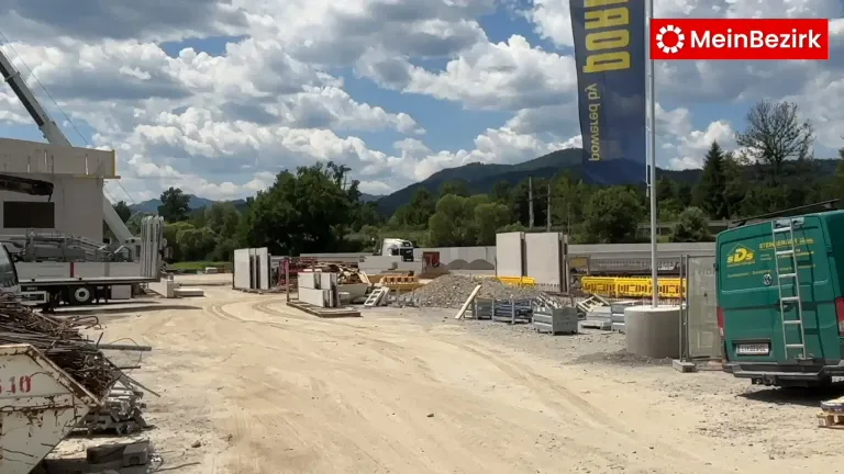 Eine aktive Baustelle mit unbefestigten Straßen, verstreuten Baumaterialien und einem Banner mit der Aufschrift "BOWEL & Co." vor dem Hintergrund von bewölktem Himmel und Bergen.