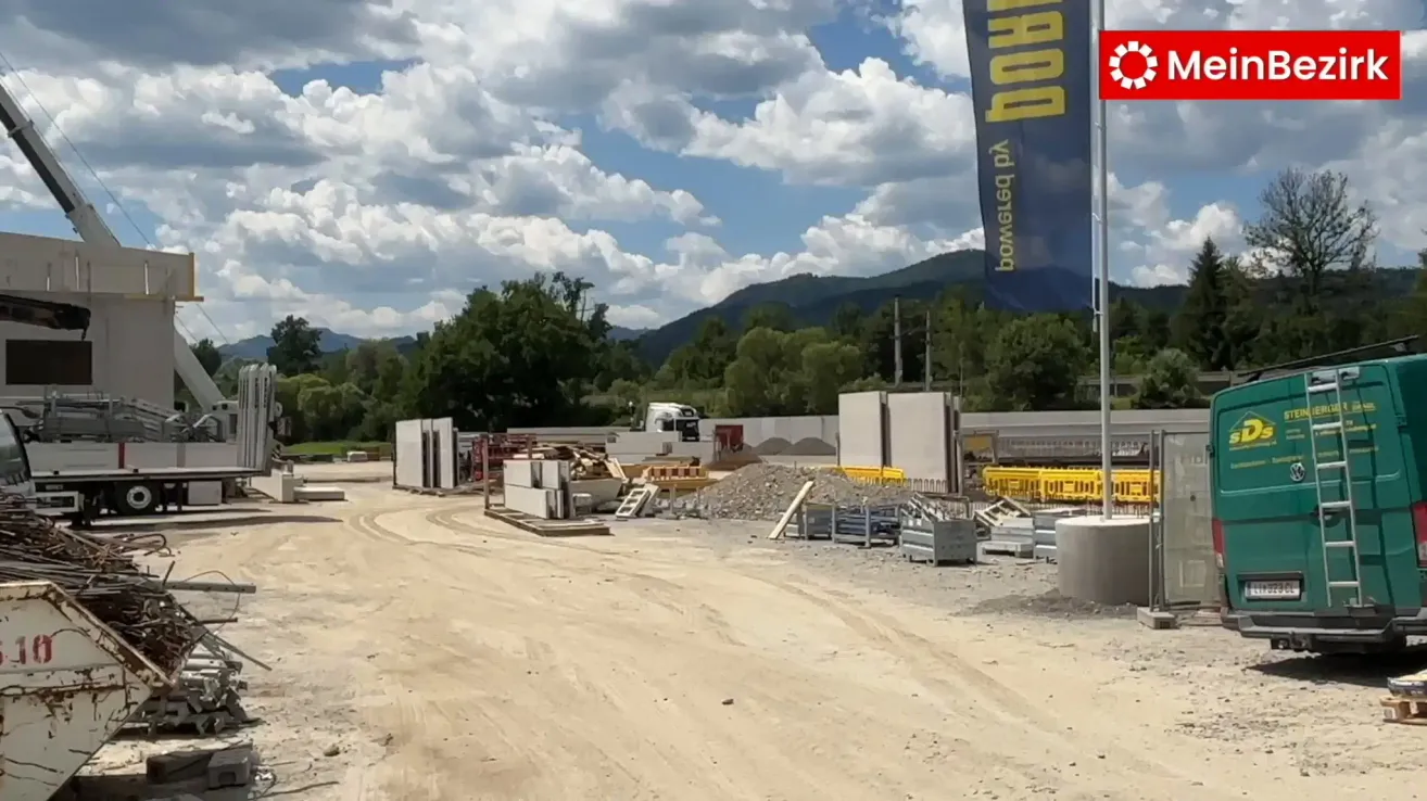 Eine aktive Baustelle mit unbefestigten Straßen, verstreuten Baumaterialien und einem Banner mit der Aufschrift "BOWEL & Co." vor dem Hintergrund von bewölktem Himmel und Bergen.