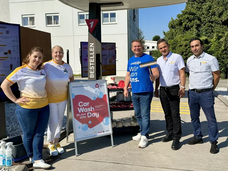Fünf Menschen posieren für ein Foto vor einer Tankstelle. Ein Schild am Boden sagt Car Wash Day. Die Männer tragen Hemden und Schuhe, die Frauen Turnschuhe und Kleider.