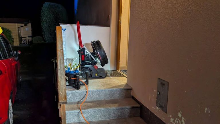 Ein roter und schwarzer Staubsauger steht neben einer Tür auf den Stufen. Ein Kabel ist sichtbar. Ein kleiner Topf und andere Gegenstände befinden sich auf dem Tisch.