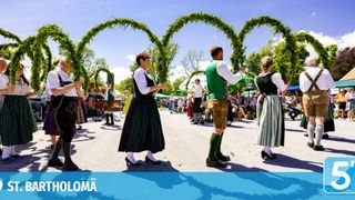 Traditionell gekleidete Personen tanzen unter dekorativen Bögen bei einem Open-Air-Festival. Sie sind von einer Menge Zuschauer umgeben.