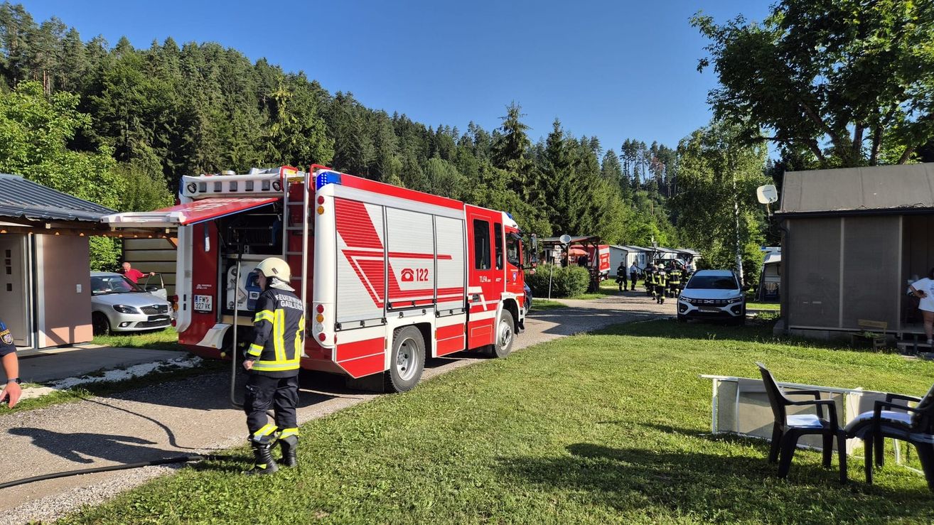 Ein Feuerwehrauto ist auf einer Straße geparkt, vor dem ein Feuerwehrmann steht. Mehrere Feuerwehrleute gehen in der Nähe. Ein weißes Auto ist auf der Seite geparkt. Bäume und Häuser sind im Hintergrund.