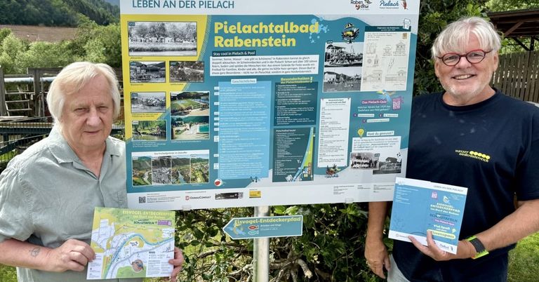Ein Schild zeigt Informationen über Pielachtalbad Rabenstein, ein historisches Spa. Zwei Personen, eine mit einer Karte und die andere mit einer Broschüre, stehen neben dem Schild.