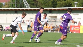 Ein Fußballspiel mit Spielern in lila und weißen Trikots, die auf einem Feld spielen. Ein Spieler steht kurz davor, den Ball zu schießen.