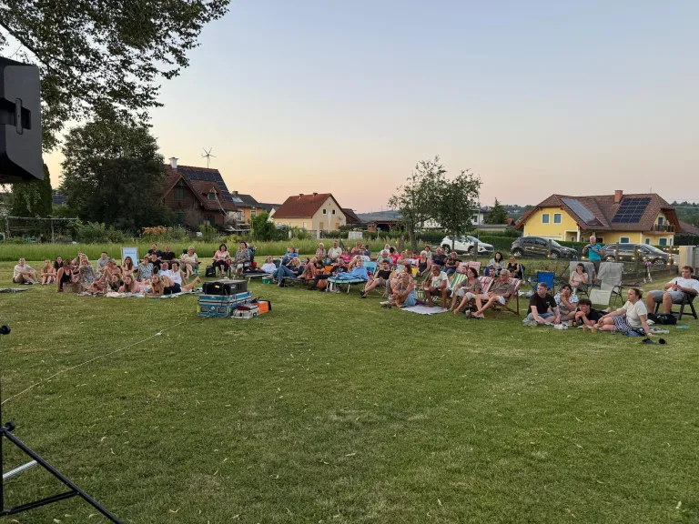 Eine Gruppe von Menschen sitzt auf einer Wiese in einer Nachbarschaft, wahrscheinlich beim Anschauen eines Films. Autos und Häuser sind im Hintergrund zu sehen.