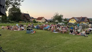 Eine Gruppe von Menschen sitzt auf einer Wiese in einer Nachbarschaft, wahrscheinlich beim Anschauen eines Films. Autos und Häuser sind im Hintergrund zu sehen.