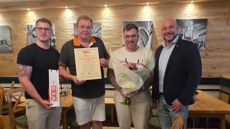 Eine Gruppe von Männern steht in einem Raum, einer hält ein Zertifikat und einen Blumenstrauß. Der Mann mit dem Strauß trägt ein weißes Hemd. Hinter ihnen hält ein anderer Mann einen Papierbeutel. Im Hintergrund befinden sich eine Holzwand und Gemälde.