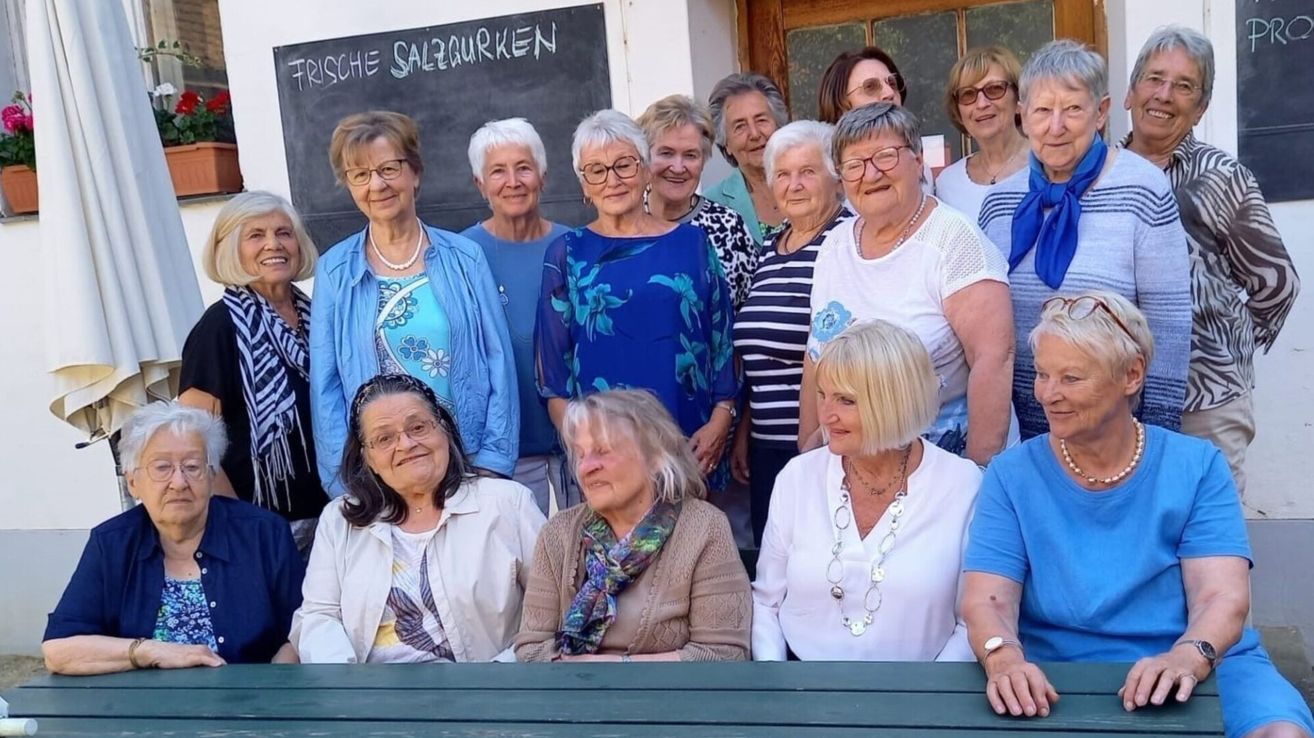 Eine Gruppe älterer Frauen steht zusammen vor einer Tafel mit 'Frische Salzgurken' darauf geschrieben. Einige tragen Brillen, Halsketten und Armbänder und lächeln für ein Foto.