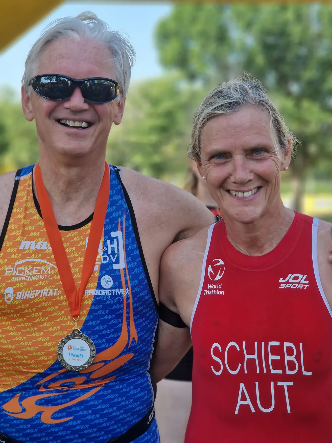 Ein lächelndes älteres Paar steht nebeneinander und trägt Triathlon-Ausrüstung. Der Mann trägt eine Medaille um den Hals und die Frau trägt ein rotes Shirt. Bäume sind im Hintergrund.