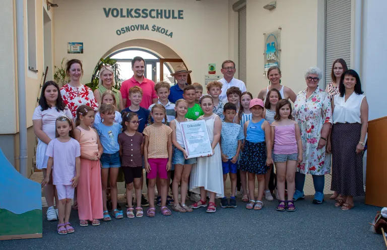 Eine Gruppe von Kindern und Erwachsenen steht vor einem Gebäude mit dem Text 'Volksschule'. Sie halten eine Urkunde in der Hand.