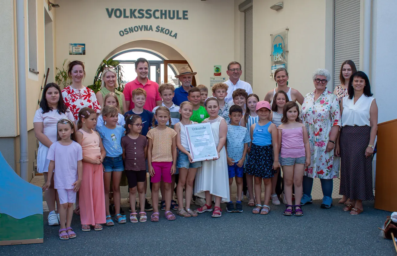Eine Gruppe von Kindern und Erwachsenen steht vor einem Gebäude mit dem Text 'Volksschule'. Sie halten eine Urkunde in der Hand.