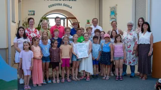 Eine Gruppe von Kindern und Erwachsenen steht vor einem Gebäude mit dem Text 'Volksschule'. Sie halten eine Urkunde in der Hand.