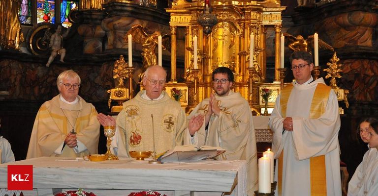 Drei Männer in religiöser Kleidung stehen vor einem Altar. Die zentrale Figur trägt ein goldenes Kreuz und hält ein offenes Buch. Zwei Kerzen auf dem Altar sind entzündet.