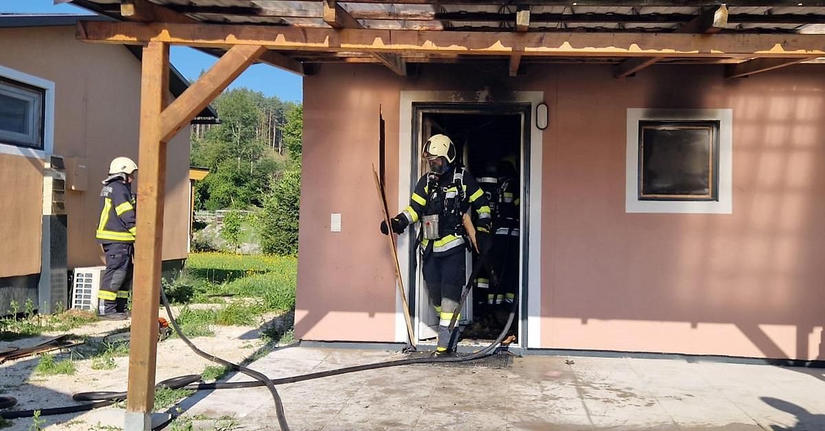 Ein Feuerwehrmann hält einen Schlauch, während ein anderer Feuerwehrmann in einem verbrannten Haus mit einer zerbrochenen Tür steht. Bäume und ein Gebäude sind im Hintergrund.