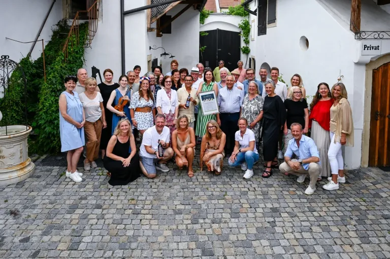 Eine Gruppe von Menschen, wahrscheinlich Kollegen, posiert für ein Foto vor einem Gebäude. Eine Person hält ein gerahmtes Zertifikat.