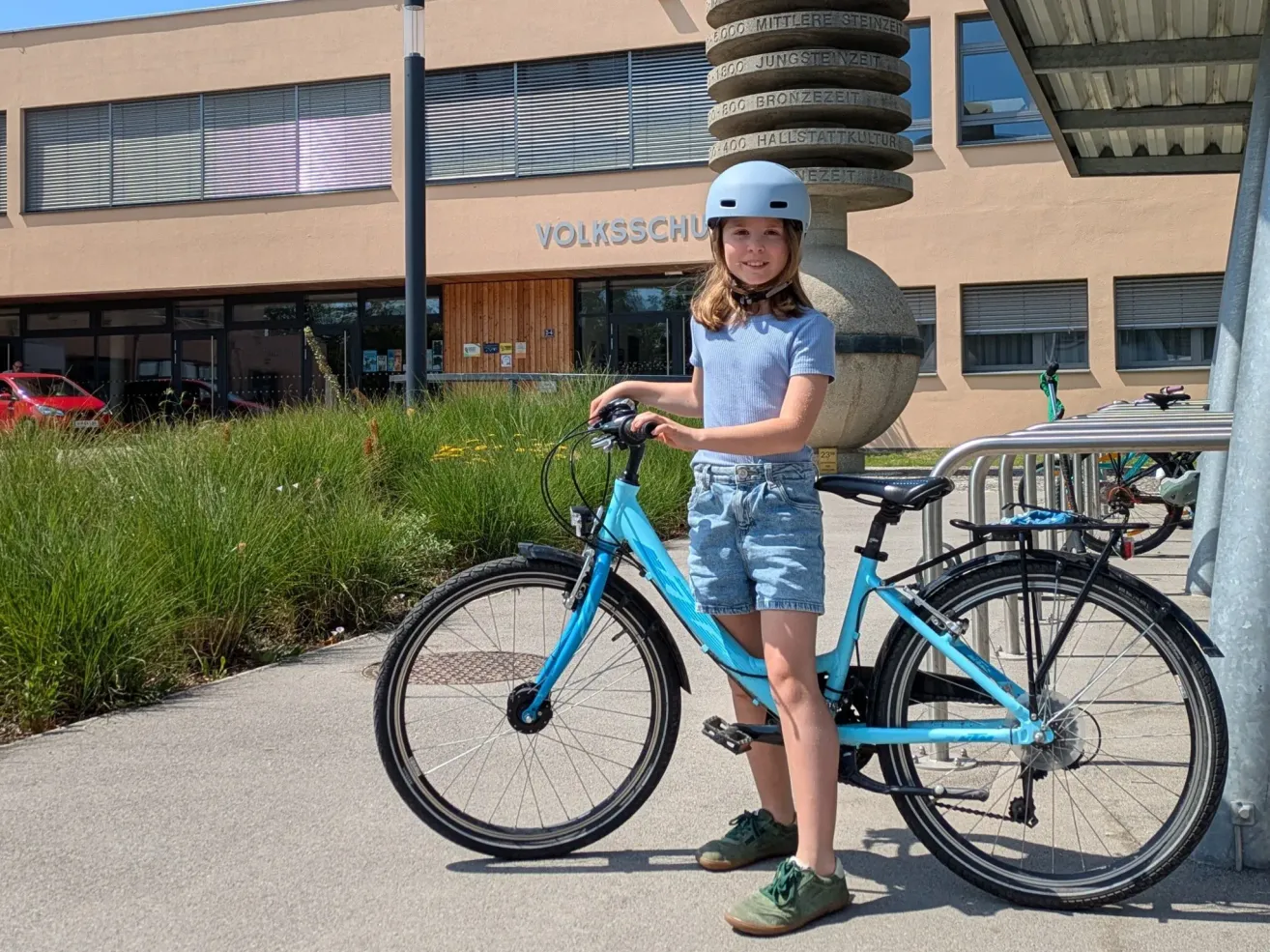 Ein Madchen mit Helm steht neben seinem blauen Fahrrad vor einem Schulgebaude.