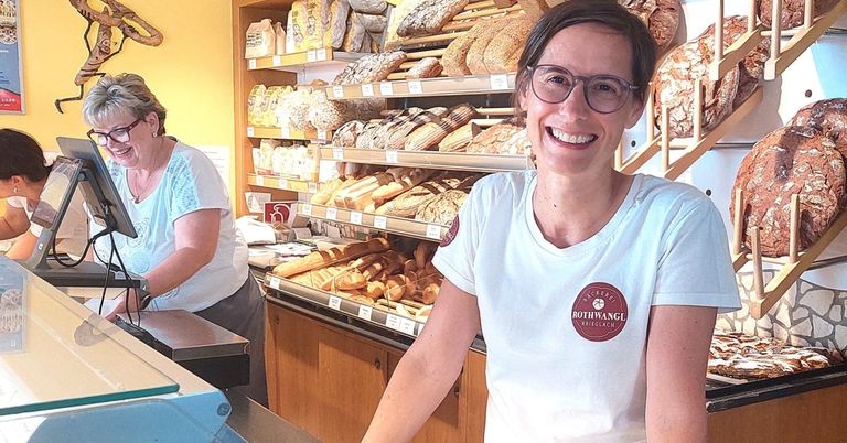 Eine lächelnde Frau mit Brille steht hinter einer Theke in einer Bäckerei mit Regalen voller Brot.
