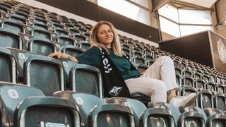 Eine Frau in einem grünen Shirt sitzt auf einem grünen Stuhl in einem Stadion mit einem schwarzen Schal mit weißen Aufschriften. Das Stadion hat viele leere Stühle und Fenster.