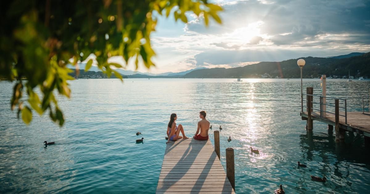 Two people sit on a wooden dock overlooking a serene lake, with mountains in the background and ducks floating nearby.