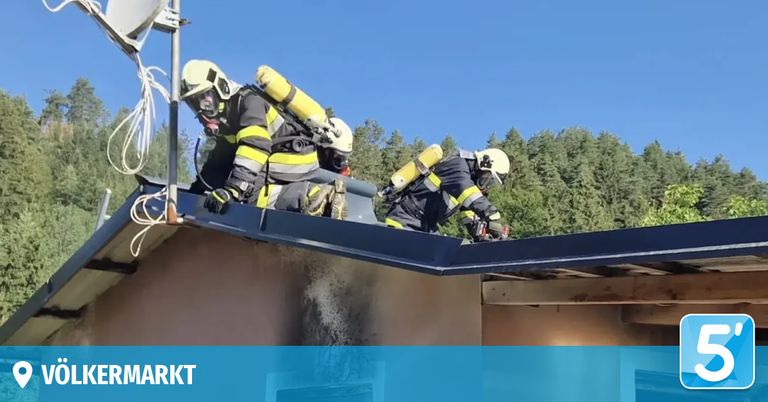 Drei Feuerwehrleute in gelben und schwarzen Uniformen mit Sauerstoffflaschen arbeiten auf einem Dach unter blauem Himmel mit Bäumen im Hintergrund.