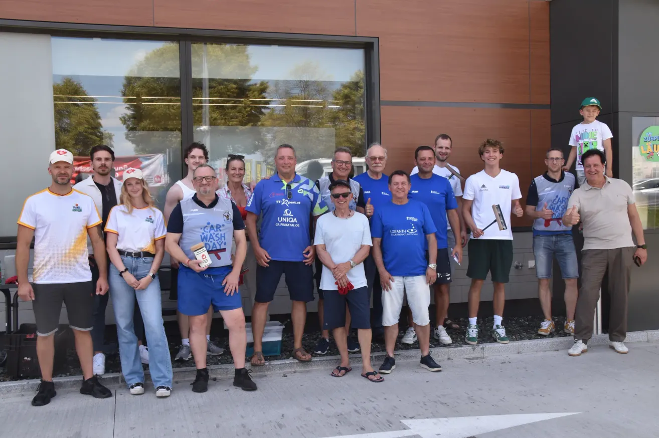 Eine Gruppe von Menschen in blauen und weißen Uniformen posiert für ein Foto vor einem Gebäude. Einige halten Tassen und Flaschen. Der Mann in der Mitte hält ein weißes Tuch. Hinter ihnen spiegelt ein Glasfenster ein Auto und Bäume.