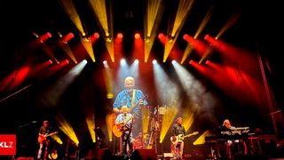 Eine Band tritt auf der Bühne mit leuchtenden roten und gelben Lichtern auf, ein Gitarrist spielt Gitarre und ein anderer Schlagzeug.