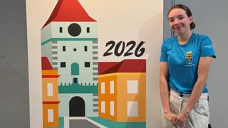 Ein lächelndes Mädchen steht neben einer Wandmalerei, die eine Stadtsilhouette mit einem Turm und Gebäuden darstellt. Das Jahr 2026 ist prominent angezeigt.
