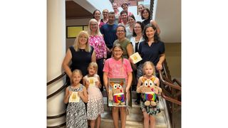 Eine Gruppe von Erwachsenen und Kindern posiert auf einer Treppe und lächelt. Zwei Mädchen halten bunte Spielzeuge und ein Mädchen eine Zeichnung. Im Hintergrund sind eine Ziegelwand und ein gerahmtes Bild zu sehen.