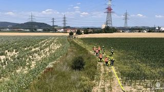 Eine Gruppe Feuerwehrleute in orangefarbenen Uniformen arbeiten in einem Feld mit einem gelben Schlauch. Dahinter befindet sich ein weites Feld mit einer Reihe von Strommasten. In der Ferne stehen Häuser und Gebäude.
