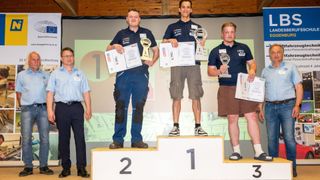 Drei Männer stehen auf einem Podium mit den Nummern 1, 2 und 3 auf den Stufen. Sie halten Trophäen und Urkunden. Ein Mann links trägt ein blaues Hemd und schwarze Hose.