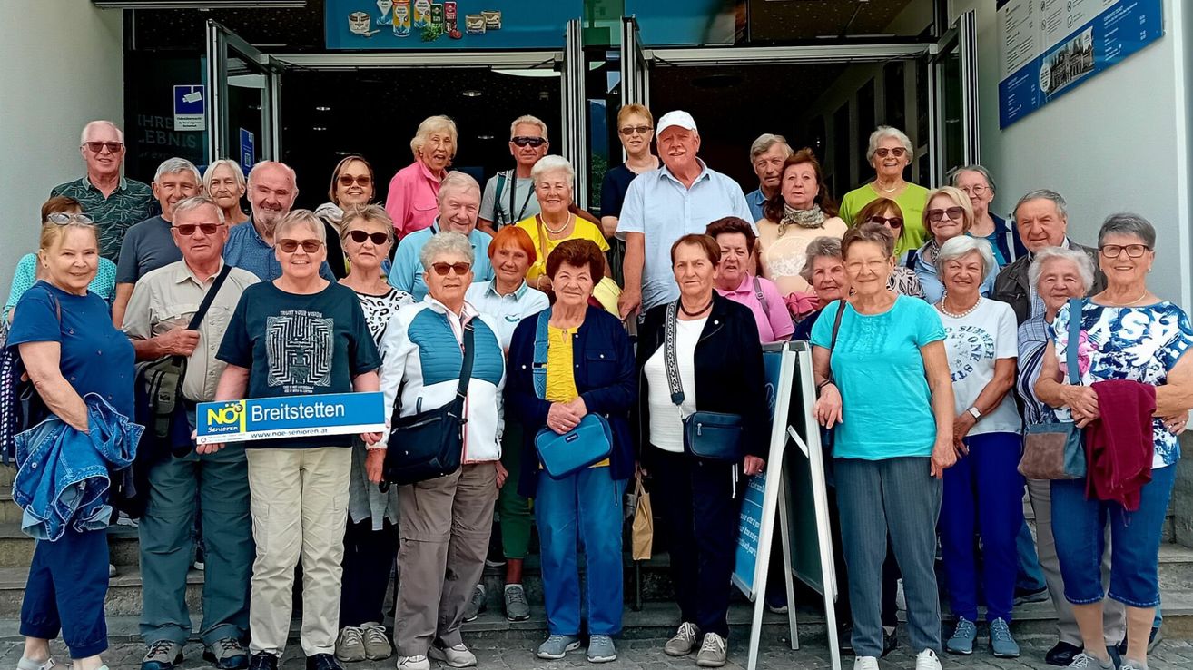 Eine Gruppe älterer Menschen posiert für ein Foto vor einem Gebäude, einige lächeln, tragen Brillen und Taschen. Einige haben Halsketten. Ein blaues Schild mit weißen Buchstaben wird von einer Person gehalten.