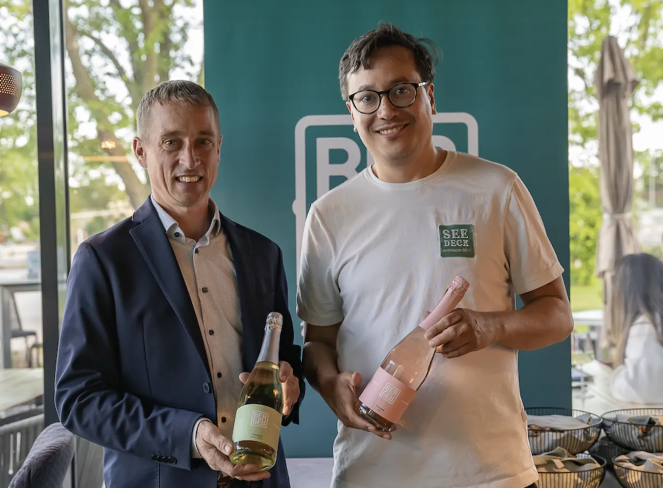 Zwei Männer stehen und lächeln, einer in Anzug hält eine Weinflasche, der andere in weißem T-Shirt mit grünem Logo hält eine ähnliche Flasche.