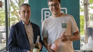 Zwei Männer stehen und lächeln, einer in Anzug hält eine Weinflasche, der andere in weißem T-Shirt mit grünem Logo hält eine ähnliche Flasche.