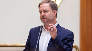 Ein Mann in einem blauen Anzug spricht in ein Mikrofon und gestikuliert mit den Händen. Er wirkt ernst, mit einer hellen Wand im Hintergrund.