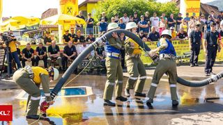 Eine Gruppe Feuerwehrleute füllt einen Pool mit Wasser, während eine Menge hinter einem Zaun zuschaut. Sie tragen Helme und halten Schläuche.