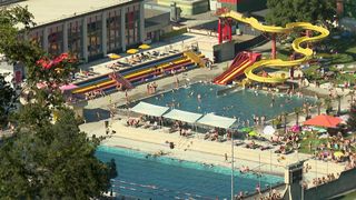 Eine Luftaufnahme eines Wasserparks zeigt zwei Schwimmbecken, zahlreiche Menschen und mehrere Wasserrutschen. Viele sind unter Sonnenschirmen oder Sonnenliegen.