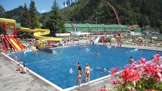 Ein Freibad mit einer gelben Wasserrutsche. Menschen schwimmen und andere liegen unter Sonnenschirmen. Ein Gebäude mit grünen Dächern ist im Hintergrund.