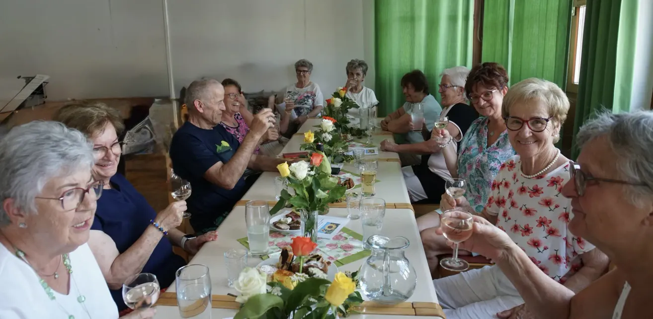 Mehrere ältere Menschen sitzen um einen Tisch mit Essen und Getränken. Eine Blumendekoration ist auf dem Tisch. Einige Leute lächeln und halten Weingläser.