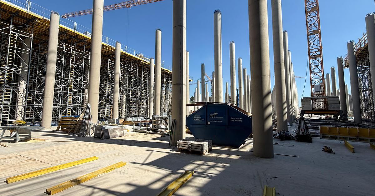 Baustelle mit großen Betonsäulen, Gerüsten und einem blauen Container mit der Aufschrift 'Service für die Zukunft'. Der Himmel ist klar und blau.