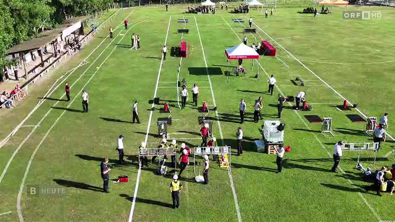 Ein Sportplatz mit mehreren Personen, einige in Uniformen, ist für eine Veranstaltung vorbereitet. Ausrüstung und Zelte sind verstreut.