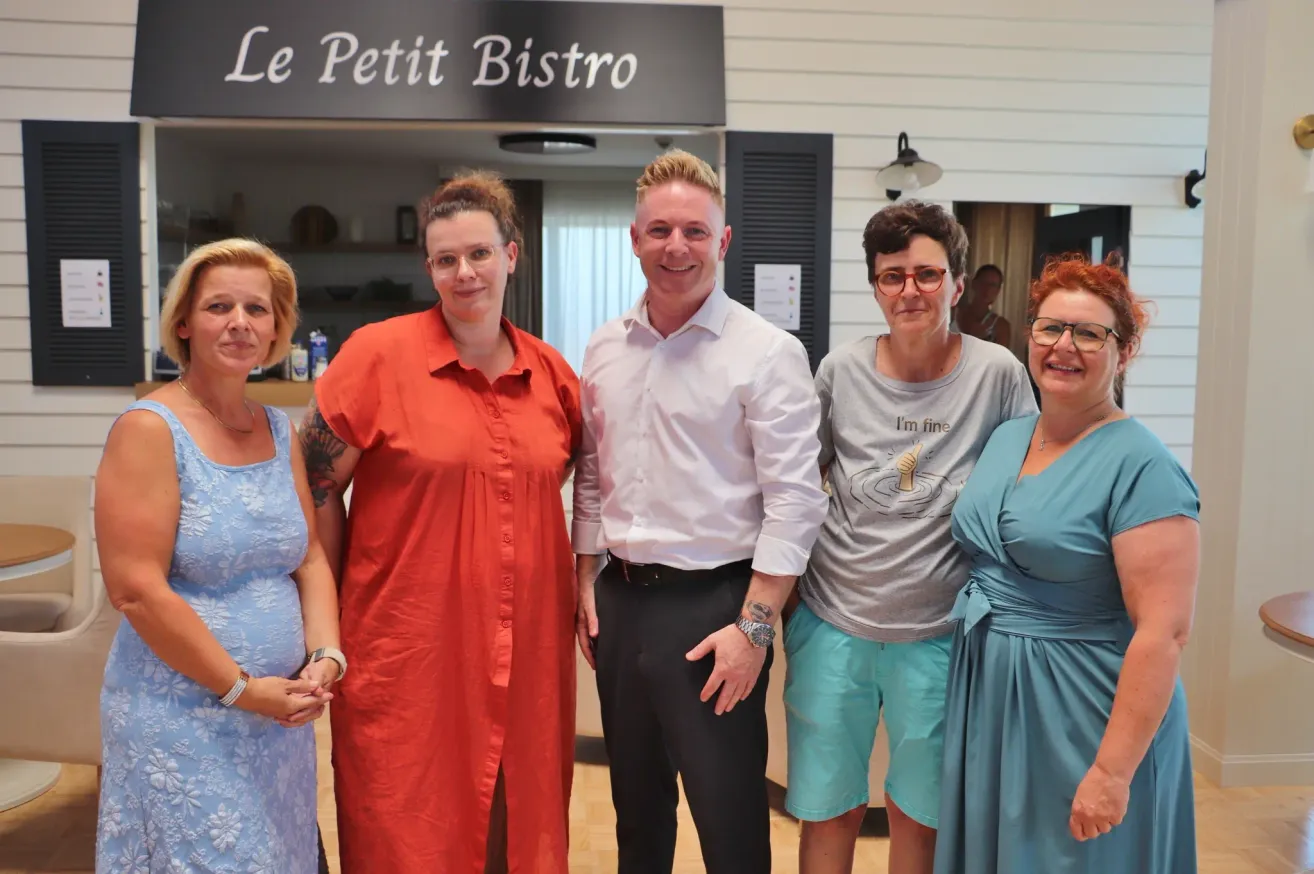 Fünf Personen posieren für ein Foto vor einem Bistro namens Le Petit Bistro. Ein Mann in einem weißen Hemd steht zwischen zwei Frauen, eine in einem orangefarbenen Shirt und die andere in einem grauen Shirt.