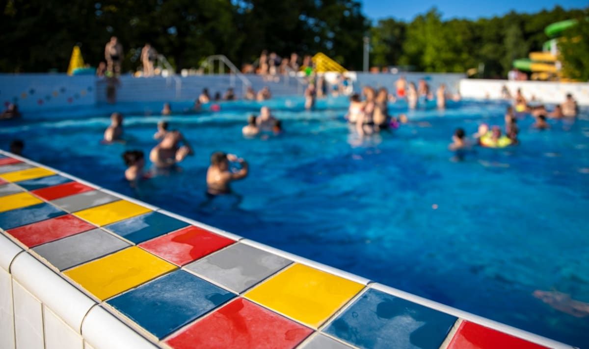 Ein überfülltes Freibad mit einem farbenfrohen, mehrfarbigen Fliesenrand. Zahlreiche Menschen schwimmen und unterhalten sich unter einem klaren Himmel.