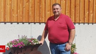 Ein Mann in einem roten Hemd und blauen Jeans steht vor einem Holzzaun mit Blumen. Er trägt eine Uhr und lächelt.