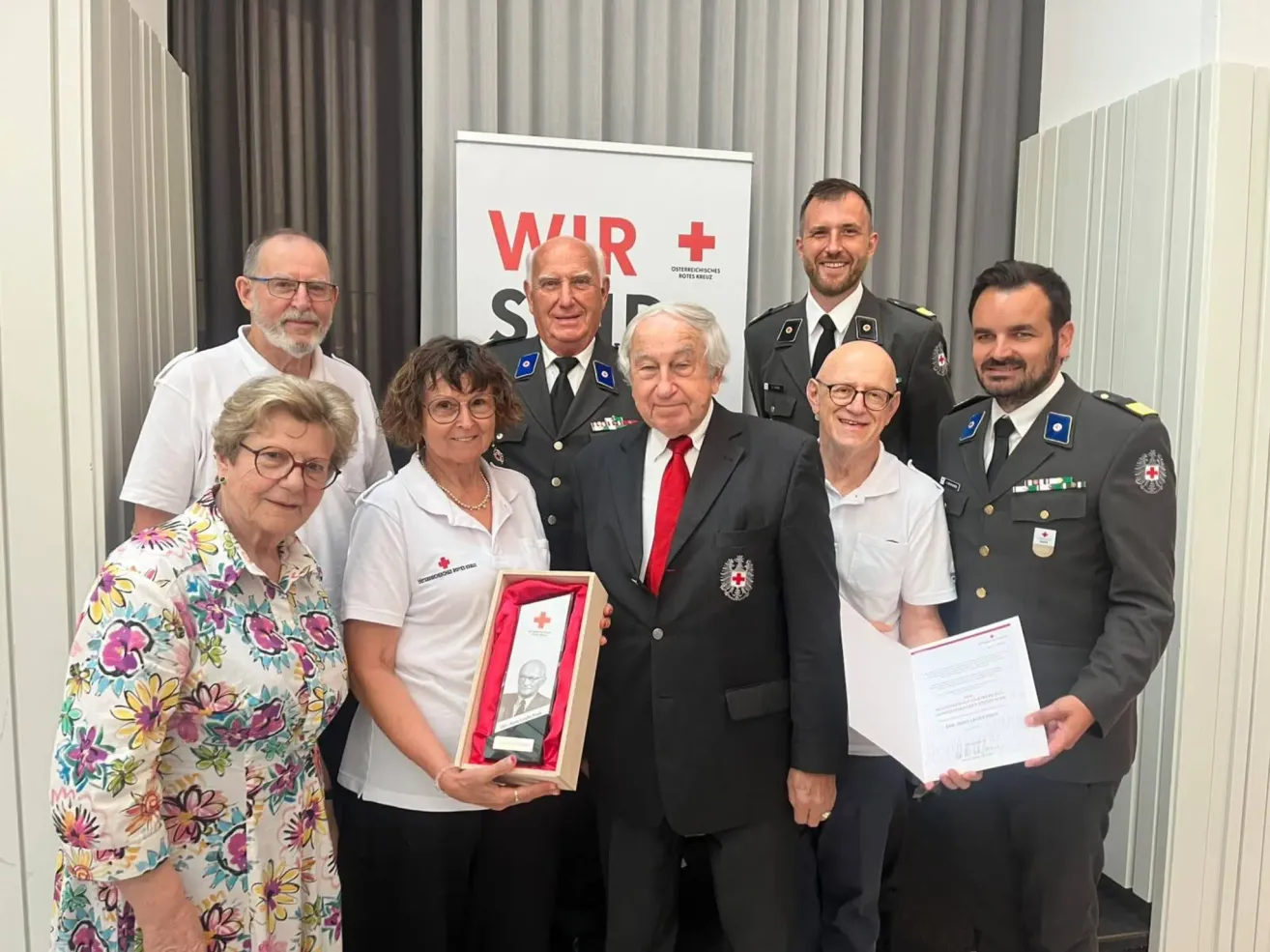 Eine Gruppe von Menschen steht gemeinsam für ein Foto. Ein Mann im Anzug hält eine gerahmte Auszeichnung. Hinter ihm halten zwei Männer in Militäruniformen Urkunden. Ein Banner in Rot und Weiß mit 'WIR +' und rotem Kreuz.