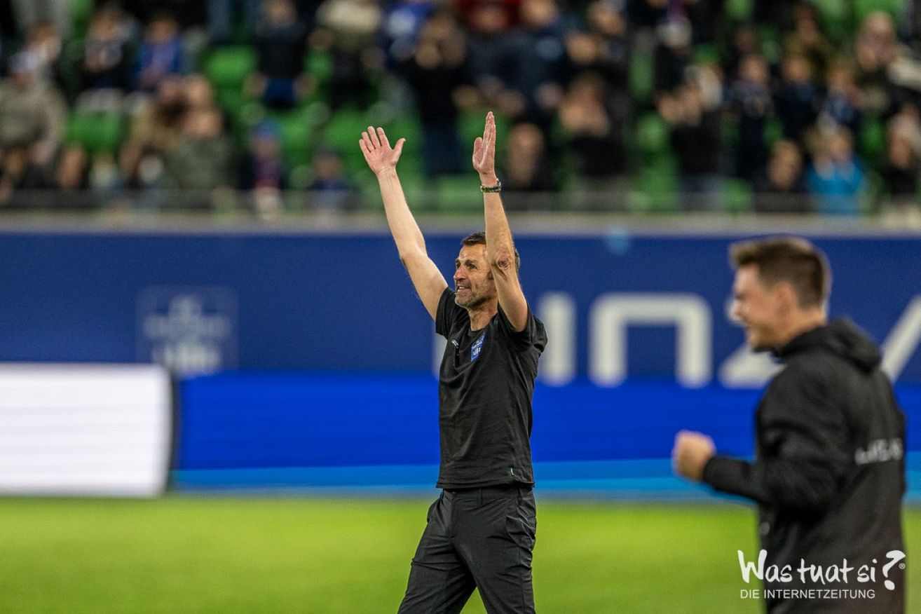Ein Fußballtrainer feiert auf dem Feld, trägt ein schwarzes Shirt und Hose. Eine blaue Wand mit Text steht hinter ihm.